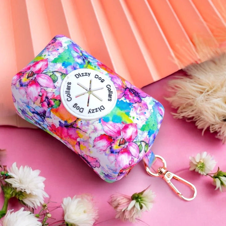 The image features a vibrant floral dog accessory bag on a pink and peach background. The bag is labeled "Dizzy Dog Collars," and the professional composition highlights the colorful design, complemented by surrounding flowers and soft textures.