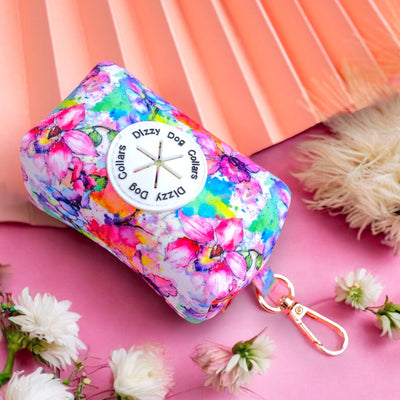 The image features a vibrant floral dog accessory bag on a pink and peach background. The bag is labeled "Dizzy Dog Collars," and the professional composition highlights the colorful design, complemented by surrounding flowers and soft textures.