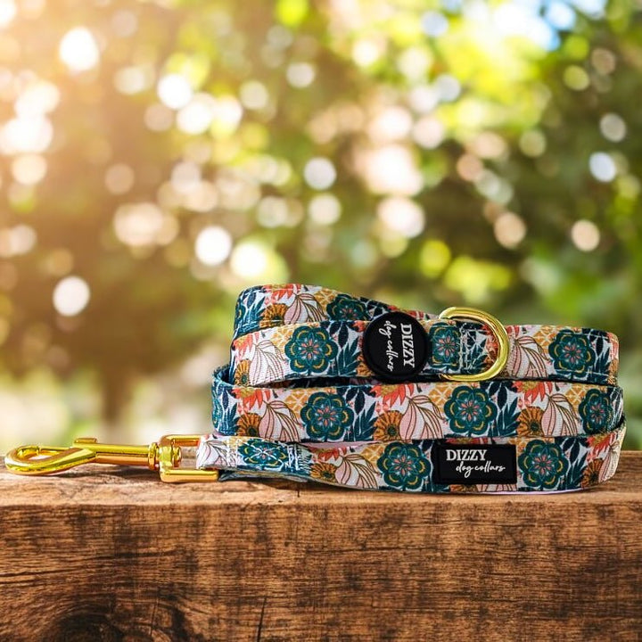 A vibrant dog leash with a floral pattern featuring green and orange flowers, and a golden clasp, laid out on a wooden surface. The background shows a soft, bokeh effect with natural light filtering through trees.