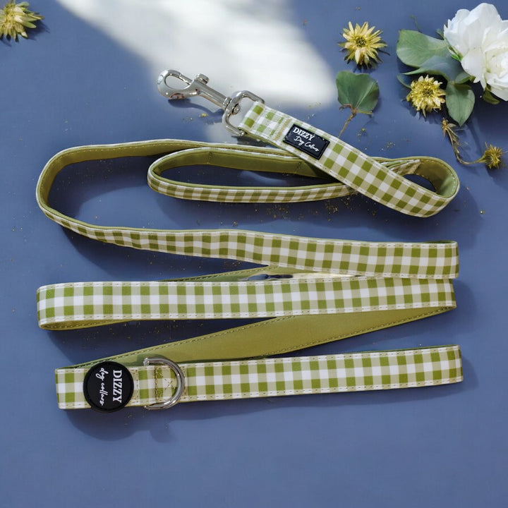 The image showcases a green and white checkered dog leash on a navy blue background, highlighted by white flowers and gold accents. The leash is labeled "Dizzy Dog Collars," and the professional photography emphasizes the leash with a decorative floral setting.