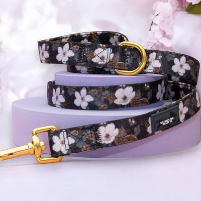 The image shows a dog leash with a floral pattern featuring white flowers against a dark background. The leash is elegantly coiled, displaying a gold metal clasp at one end and a gold D-ring at the other. The leash is set on a light purple platform, with soft pink cherry blossoms in the background, enhancing its overall elegant and stylish look. A small black label with white text adds a touch of branding to the product.