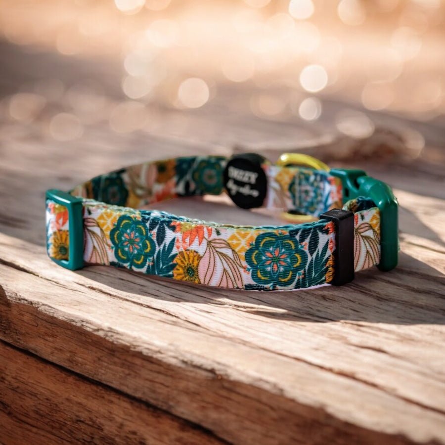 A colourful dog collar with a floral pattern featuring green and orange flowers, and a green buckle, displayed on a wooden surface. The background has a soft, bokeh effect with natural light filtering through trees.