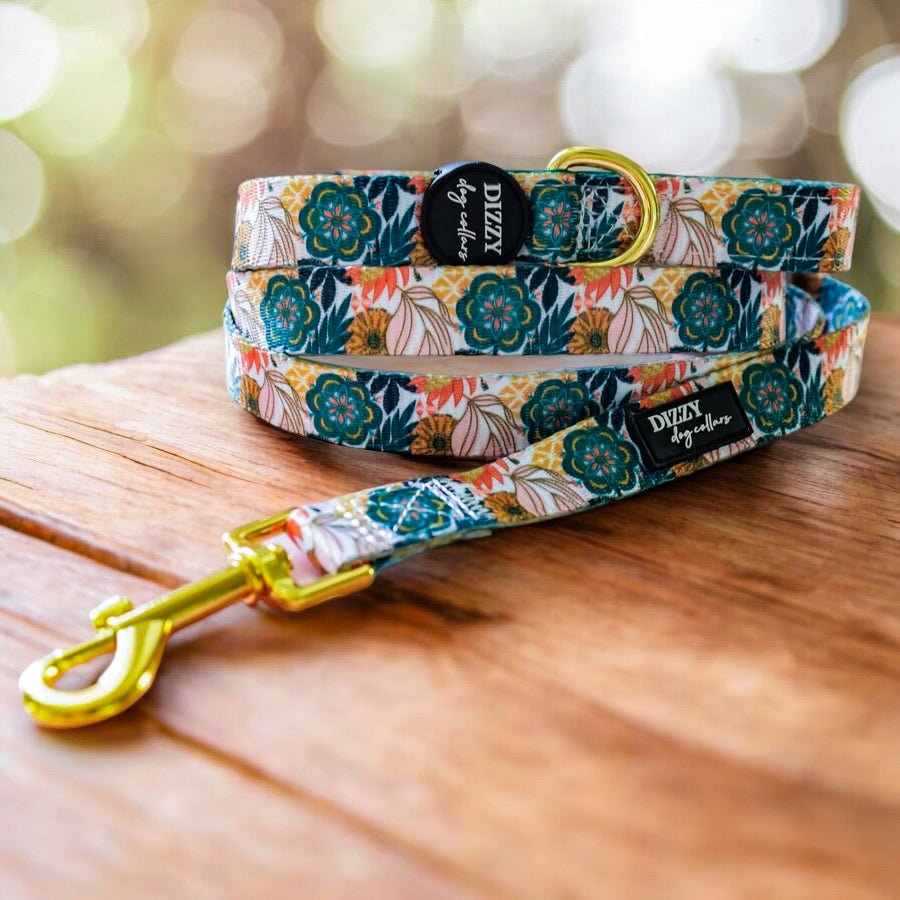 A colourful dog leash with a floral pattern featuring green and orange flowers, and a golden clasp, coiled on a wooden surface. The background is filled with sunlight filtering through a lush, green environment