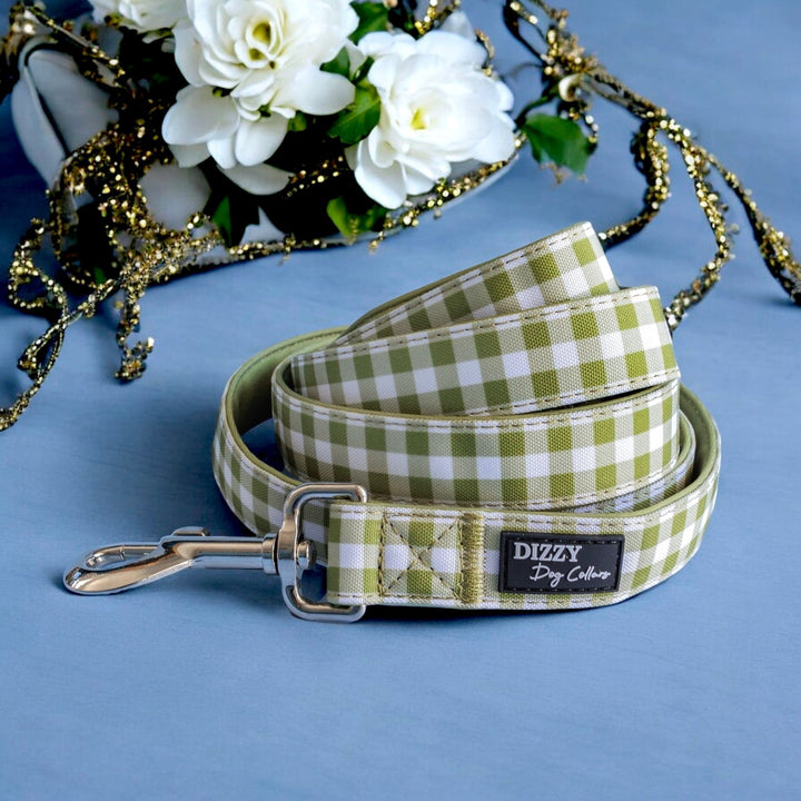 The image showcases a green and white checkered dog leash neatly coiled on a navy blue background, adorned with white roses and gold decorations. The leash is labeled "Dizzy Dog Collars," and the arrangement emphasizes the leash with a sophisticated floral backdrop.