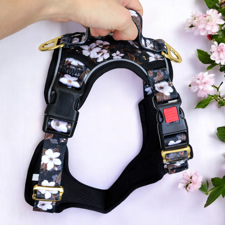 This image features a close-up of a hand holding a black dog harness with a floral pattern. The design includes white flowers with brown and green accents on a dark background, similar to the previous harness. The harness has gold hardware, including buckles and D-rings, with a black quick-release buckle that has a red locking mechanism for added security. The harness is placed against a light pink background, and there are some pink flowers on the right side of the image.