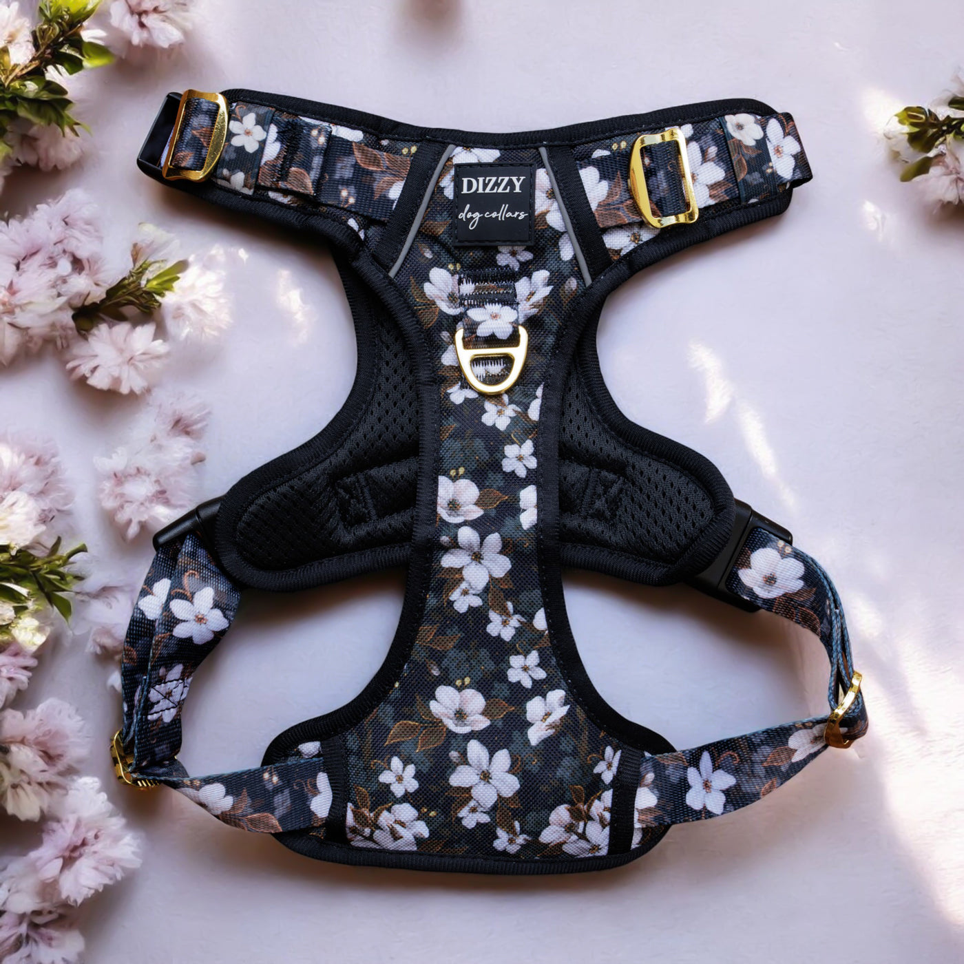 This image shows a black dog harness with a floral pattern. The design features white flowers with subtle brown and green accents on a dark background. The harness has gold hardware, including buckles and a D-ring, and is well-padded for comfort. The brand name "DIZZY dog collars" is visible on a label stitched onto the harness. The harness is laid out on a light grey surface, with some small white flowers on the left side of the image.