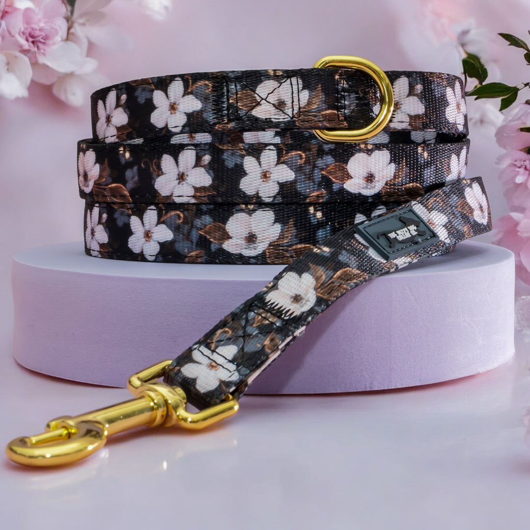 The image showcases a dog leash with a floral pattern featuring white flowers against a dark background. The leash is neatly coiled with a gold metal clasp at one end and a gold D-ring. It is displayed on a light purple platform, with soft pink cherry blossoms in the background, enhancing its elegant and stylish design. The leash also features a small black label with white text, adding a touch of branding to the product.