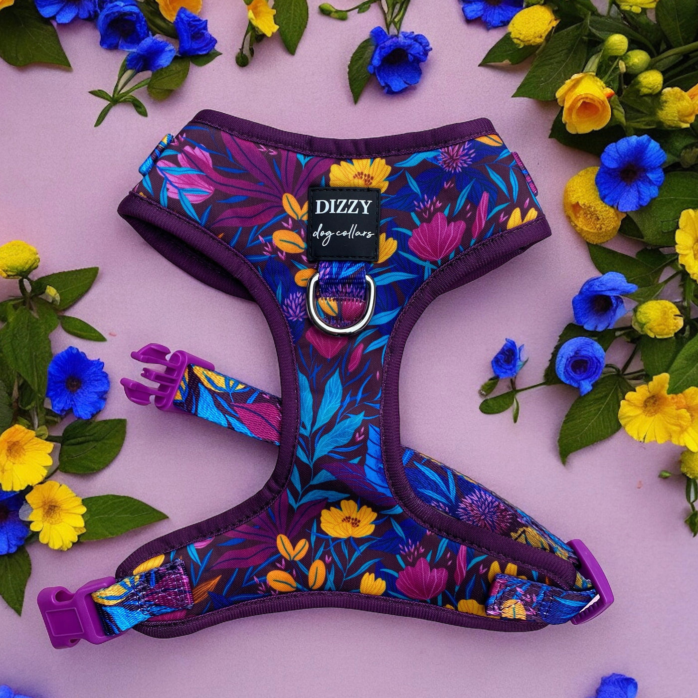 A vibrant dog harness from Dizzy Dog Collars is displayed on a pink background, surrounded by yellow and blue flowers. The harness features a colourful floral pattern with shades of purple, blue, yellow, and pink. It has purple clasps and a metal D-ring for attaching a leash. The design is lively and eye-catching, perfect for adding a touch of style to a dog's outfit.