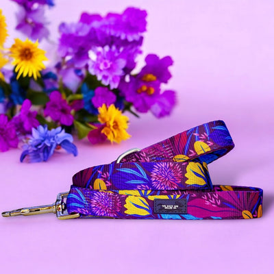 A vibrant dog leash with a colourful floral pattern, featuring a silver clasp. The leash is laid out on a soft pink background. In the background, there is a bouquet of purple, blue, and yellow flowers, adding a lively and colourful touch to the scene.