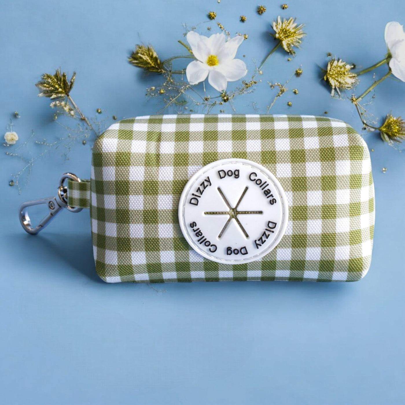 The image features a green and white checkered dog bag on a light blue background, adorned with white and gold flowers. The bag is labeled "Dizzy Dog Collars," and the composition focuses on the bag with an elegant touch of floral decorations.