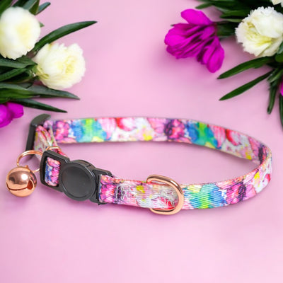 The image features a colorful floral-patterned dog collar with a rose gold bell and buckle, laid out on a pink background. The collar is surrounded by white and purple flowers, creating an elegant and visually appealing composition that highlights the vibrant design of the collar.