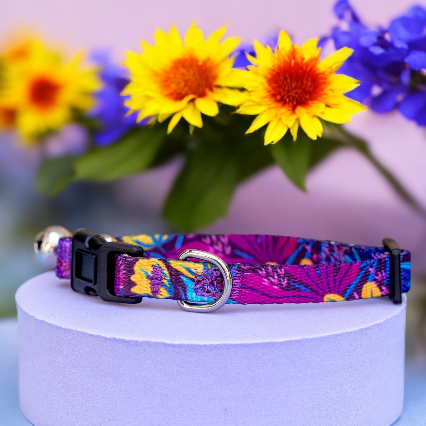 A vibrant dog collar with a colourful floral pattern, featuring a black buckle and a silver D-ring, displayed on a light purple cylindrical stand. In the background, there are bright yellow and orange flowers resembling sunflowers, along with blue flowers and green leaves, set against a soft pastel background.