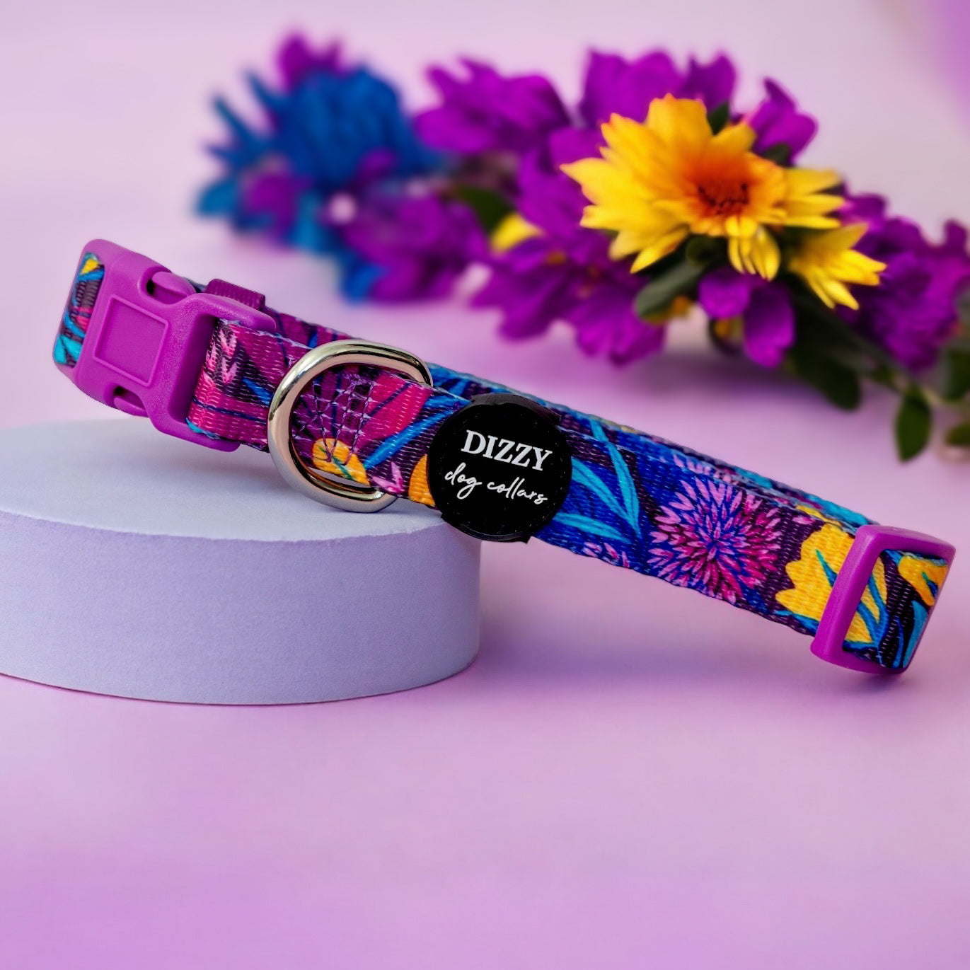 A colourful dog collar with a vibrant floral pattern, featuring a purple buckle, a silver D-ring, and a black label with "Dizzy Dog Collars" text. The collar is displayed on a light purple cylindrical stand, with a bouquet of purple, blue, and yellow flowers in the background, set against a soft pink backdrop.