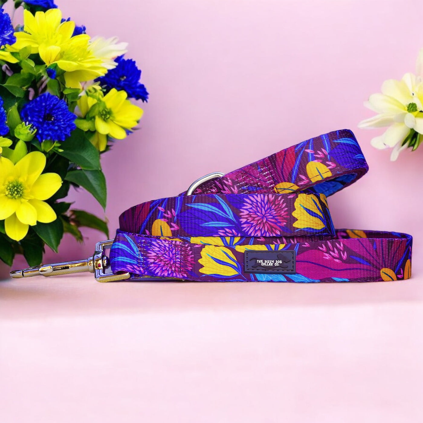 a vibrant dog leash with a colourful floral pattern, featuring a silver clasp. The leash is laid out on a soft pink background. In the background, there is a bouquet of bright yellow and blue flowers with green leaves, enhancing the lively and colourful appearance of the scene.