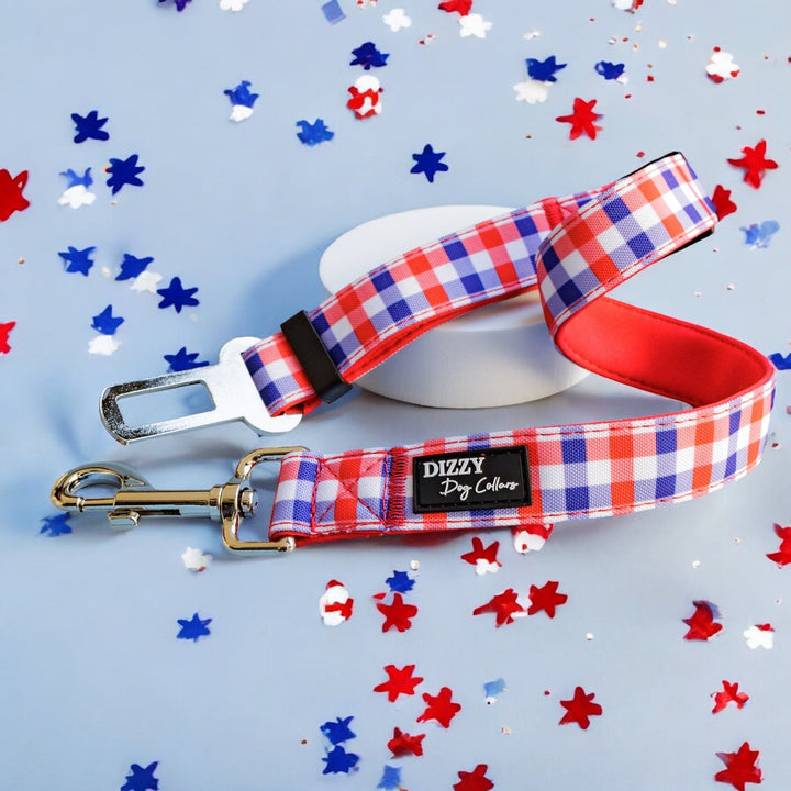 Red, white, and blue gingham-patterned dog seatbelt attachment with a silver clasp and seatbelt buckle, featuring a black Dizzy Dog Collars logo. The leash is displayed against a light blue background with scattered red, white, and blue star-shaped confetti