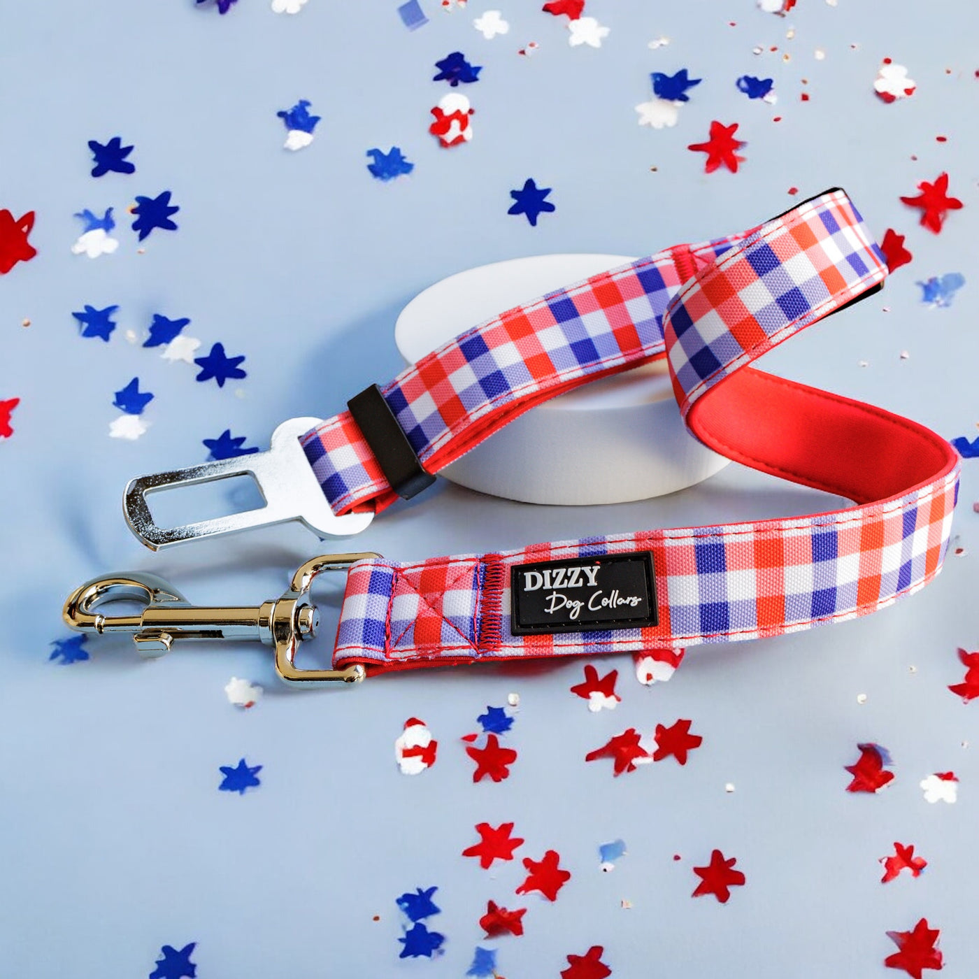 Red, white, and blue gingham-patterned dog seatbelt attachment with a silver clasp and seatbelt buckle, featuring a black Dizzy Dog Collars logo. The leash is displayed against a light blue background with scattered red, white, and blue star-shaped confetti