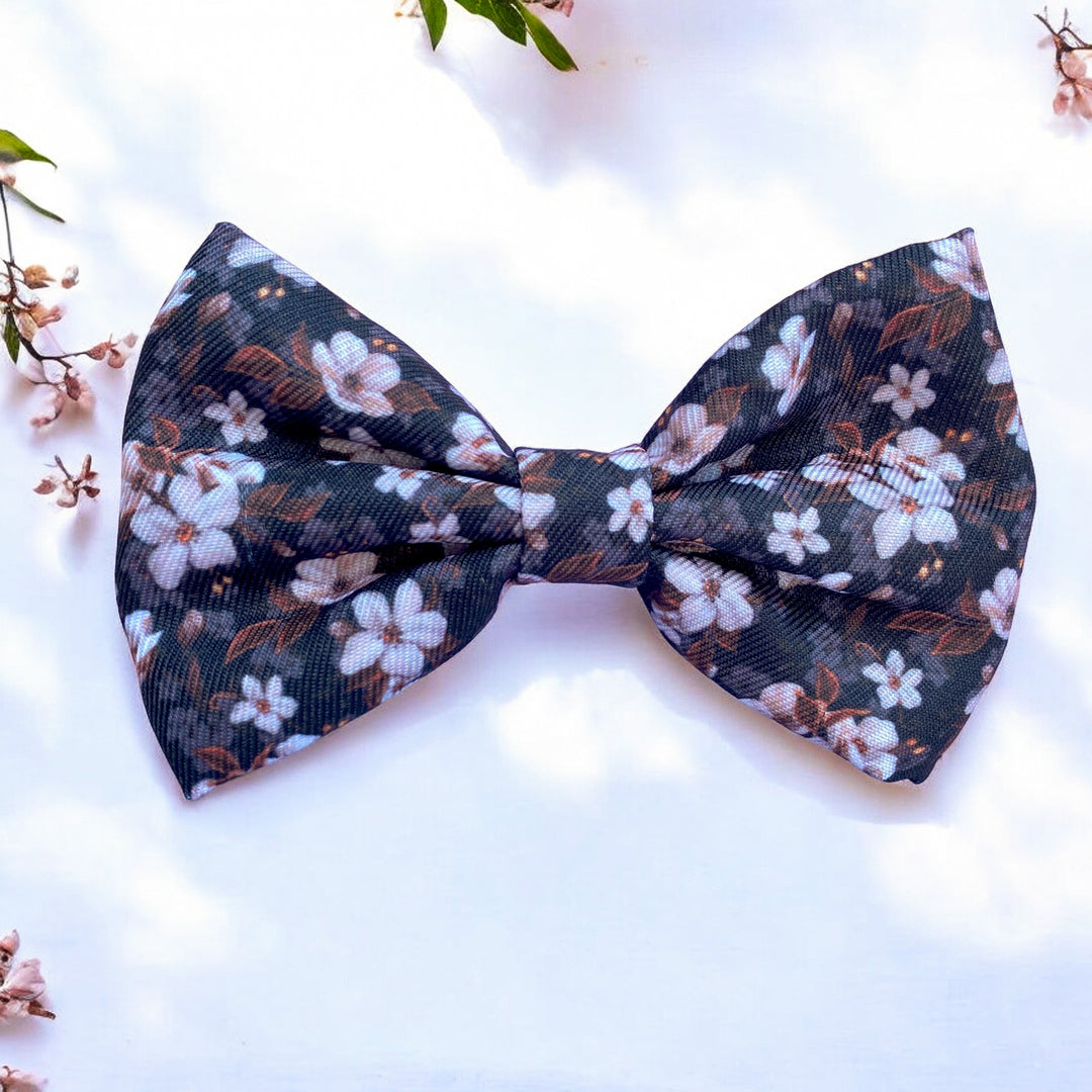 The image shows a dog bow tie with a floral pattern featuring white flowers against a dark background. The bow tie is neatly arranged on a light surface with small pink flowers surrounding it, adding a touch of elegance to the presentation. The design combines a stylish floral aesthetic with a functional accessory for dogs, making it both fashionable and practical.