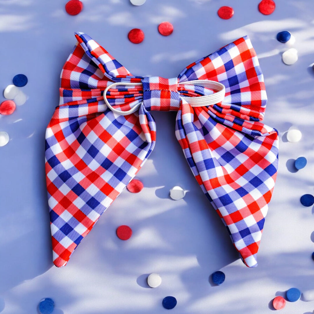 A large red, white, and blue gingham-patterned bow tie with pointed ends, displayed on a light blue background. Red and white star-shaped confetti is scattered around the bow