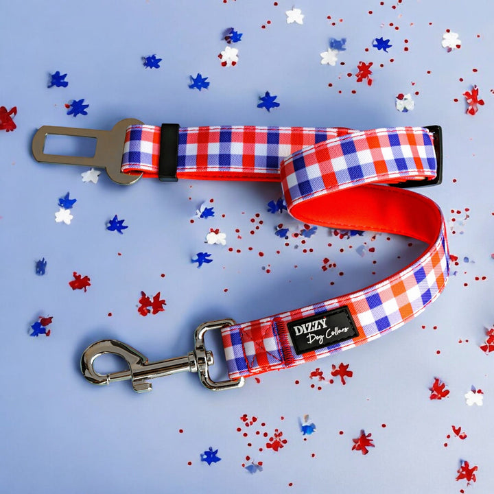 Red, white, and blue gingham-patterned dog seatbelt attachment with a silver clasp and seatbelt buckle, featuring a soft red padded underside and a black Dizzy Dog Collars logo. Displayed flat on a light blue background with scattered red, white, and blue confetti