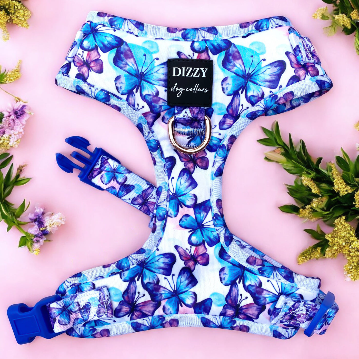 A vibrant dog harness decorated with a butterfly pattern in shades of blue and purple is displayed against a light pink background. The harness features a sturdy D-ring and adjustable blue plastic buckles. The label reads "DIZZY dog collars." The image is adorned with sprigs of flowers in green, yellow, and purple hues on the sides, adding a natural touch to the presentation.