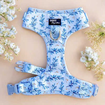 This image shows a light blue dog harness with a delicate floral pattern featuring blue leaves and orange accents. The harness is displayed flat on a soft peach background, surrounded by sprigs of white and yellow flowers, enhancing its aesthetic appeal. The design includes adjustable straps, a D-ring for leash attachment, and a branded label that reads "Dizzy Dog Collars" at the center.