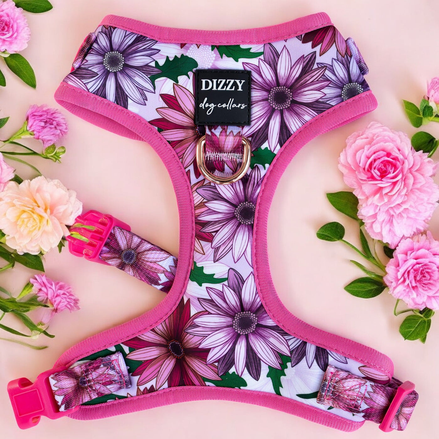 This image shows a vibrant pink dog harness adorned with a floral pattern featuring large white and purple daisies against a white background. The harness is displayed flat on a pink surface, surrounded by pink carnations and green foliage, enhancing its colorful appearance. It features a pink buckle, adjustable straps, and a branded label that reads "Dizzy Dog Collars" in the center. The design is both functional and stylish, ideal for fashionable pets.