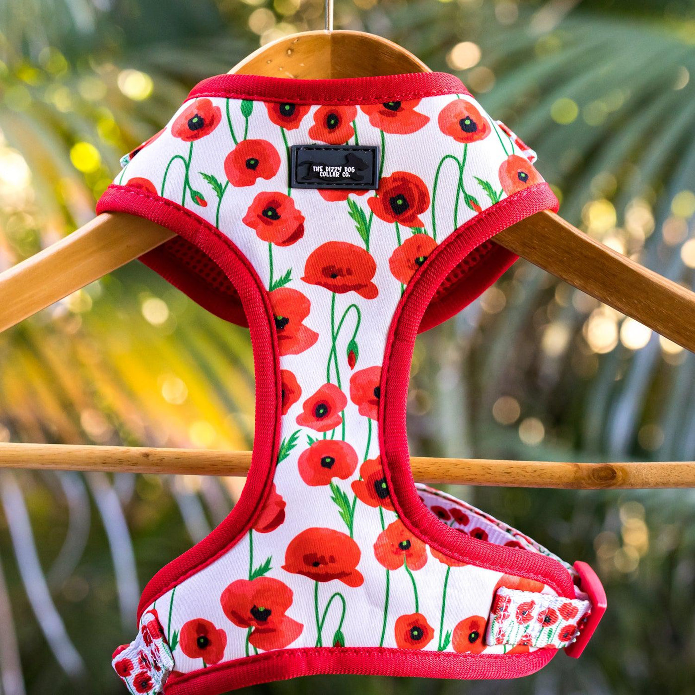 A dog harness featuring a vibrant design of red poppies with green stems on a white background is hanging on a wooden hanger. The harness has red trim around the edges and a label that reads "The Dizzy Dog Collar Co." The background shows blurred greenery, indicating an outdoor setting with a natural, sunny ambiance