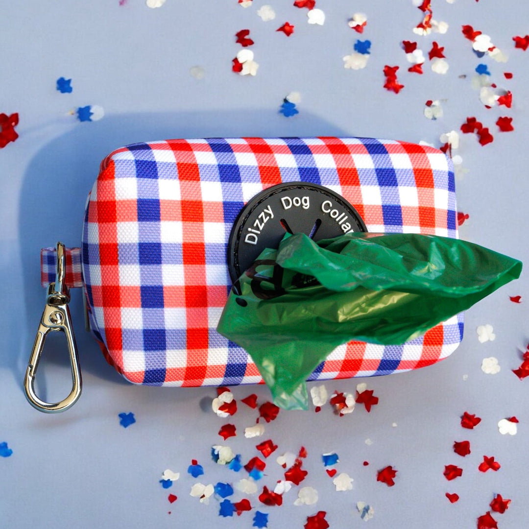 Red, white, and blue gingham-patterned waste bag holder with a green dog waste bag partially pulled out, featuring a black Dizzy Dog Collars logo and a silver clip for easy attachment. Displayed on a light blue background with scattered red, white, and blue confetti