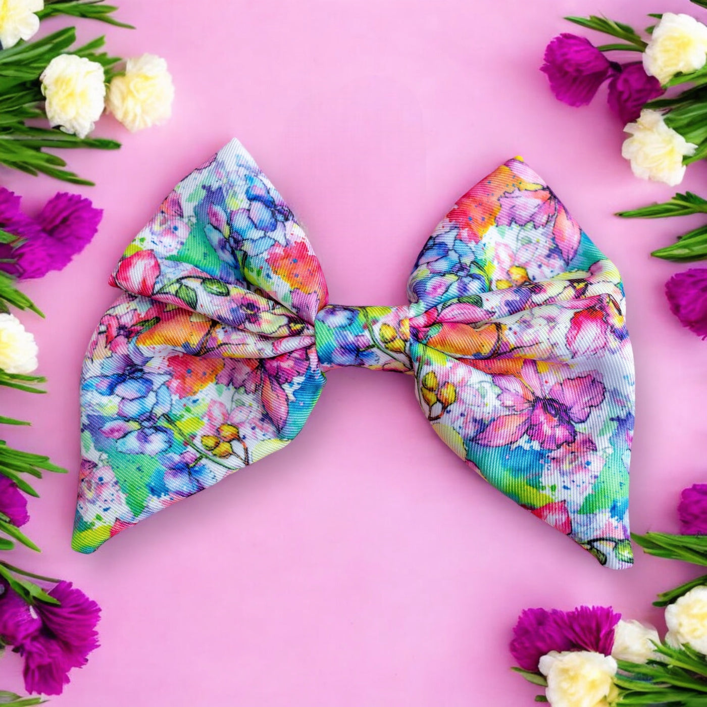 This image shows a brightly coloured bow tie with a floral watercolour design. The bow tie features vibrant shades of pink, purple, green, and blue, creating a lively and eye-catching pattern. The bow is placed on a pink background, surrounded by clusters of artificial flowers in white, purple, and yellow, enhancing the overall cheerful and vibrant aesthetic.