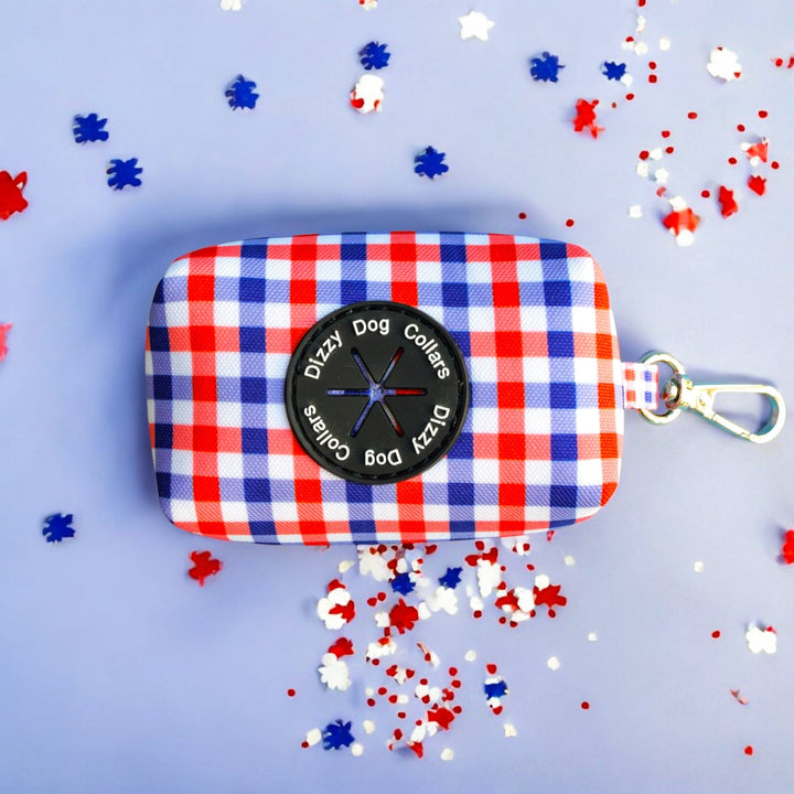 Red, white, and blue gingham-patterned waste bag holder with a black Dizzy Dog Collars logo and a silver clip for easy attachment. Displayed flat on a light blue background, surrounded by scattered red, white, and blue confetti