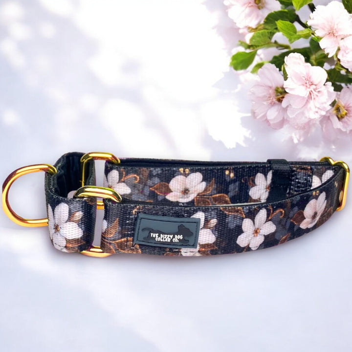 The image features a martingale dog collar with a floral pattern of white flowers against a dark background. The collar is displayed on a light surface with blooming pink cherry blossoms in the background, enhancing its elegant appearance. The collar has gold metal rings and a black label with white text that reads "The Dizzy Dog Collars Co." The combination of the floral design and the high-quality hardware gives the collar a stylish and functional look.