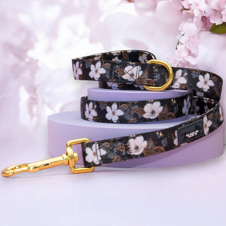 The image features a dog leash with a floral pattern of white flowers against a dark background. The leash is coiled neatly and showcases a gold metal clasp at one end and a gold D-ring. The leash is displayed on a light purple platform with soft pink cherry blossoms in the background, enhancing the overall elegant and stylish design.