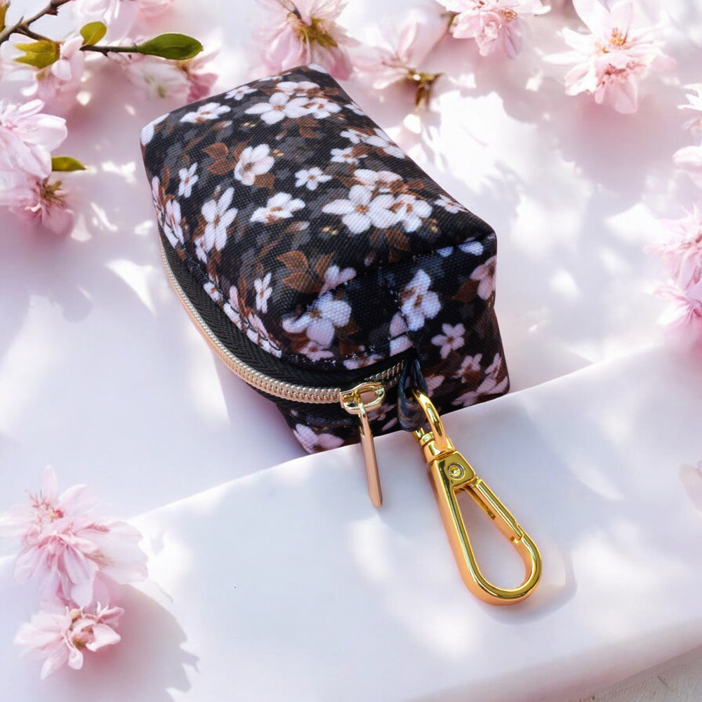 The image shows a waste bag holder with a pattern of white flowers against a dark background. The holder is rectangular with a zippered top and a gold metal clip for attaching to a leash or belt. The holder is placed on a light surface with pink cherry blossoms around it, creating a soft and elegant setting. The design combines practicality with a floral aesthetic.