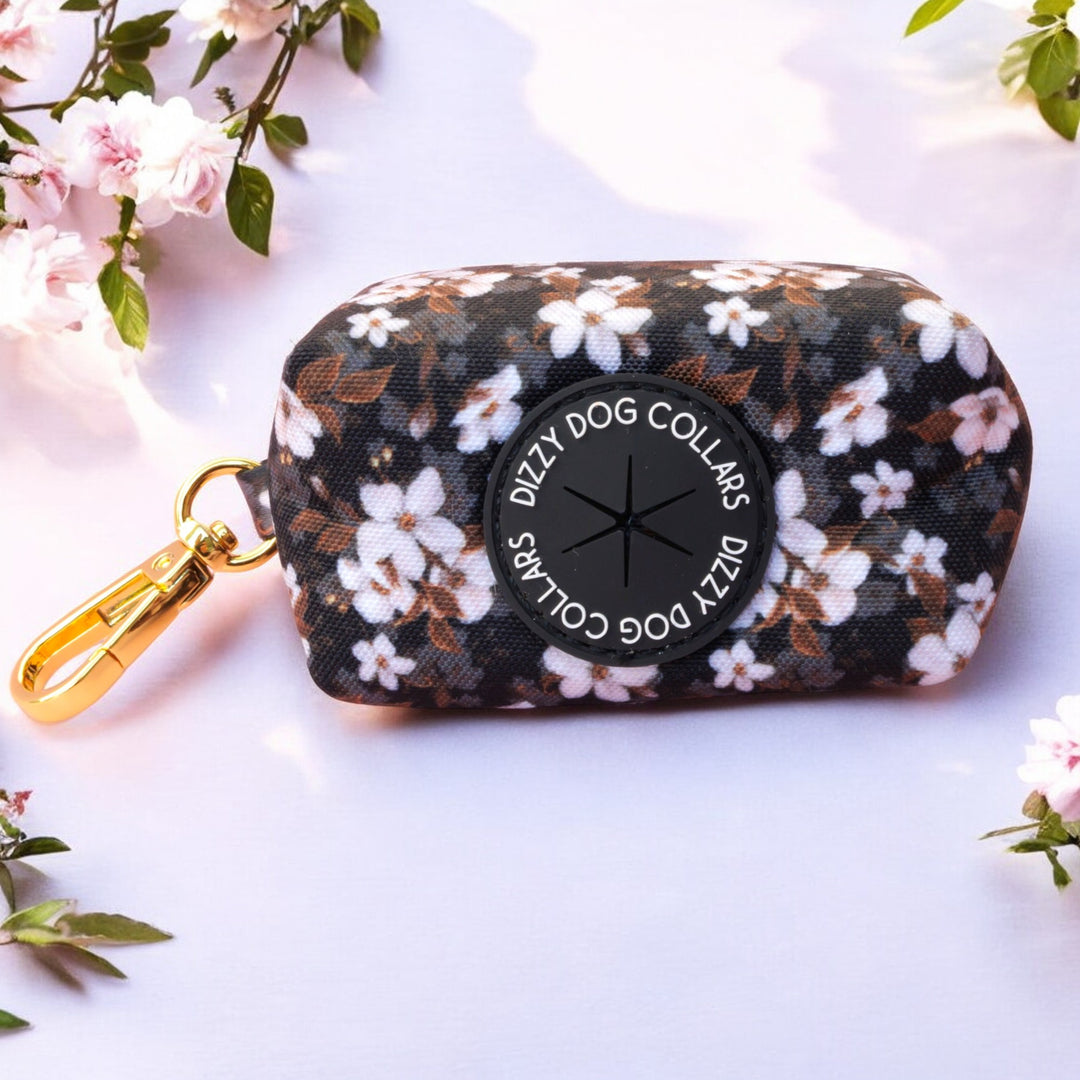 The image shows a waste bag holder featuring a pattern of white flowers against a dark background. The holder has a black circular opening labeled "Dizzy Dog Collars" for dispensing bags. It includes a gold metal clip for attaching to a leash or belt, combining functionality with a stylish floral design. The background includes soft pink cherry blossoms, adding a touch of elegance.