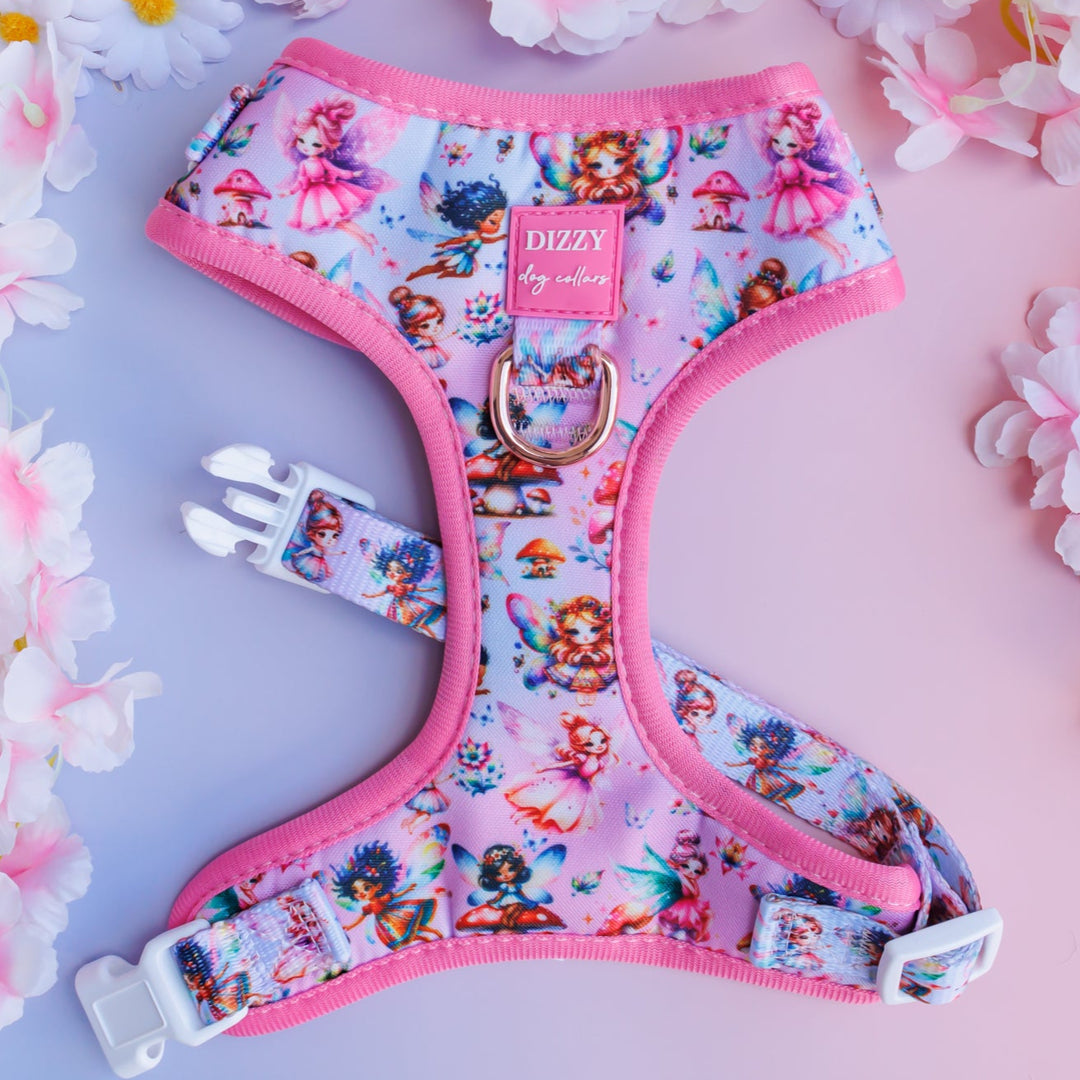 Pink dog harness with a whimsical fairy-themed design, featuring colourful illustrations of fairies, mushrooms, and flowers. The harness has pink trim and white buckles, displayed on a pastel backdrop with white and pink floral accents.