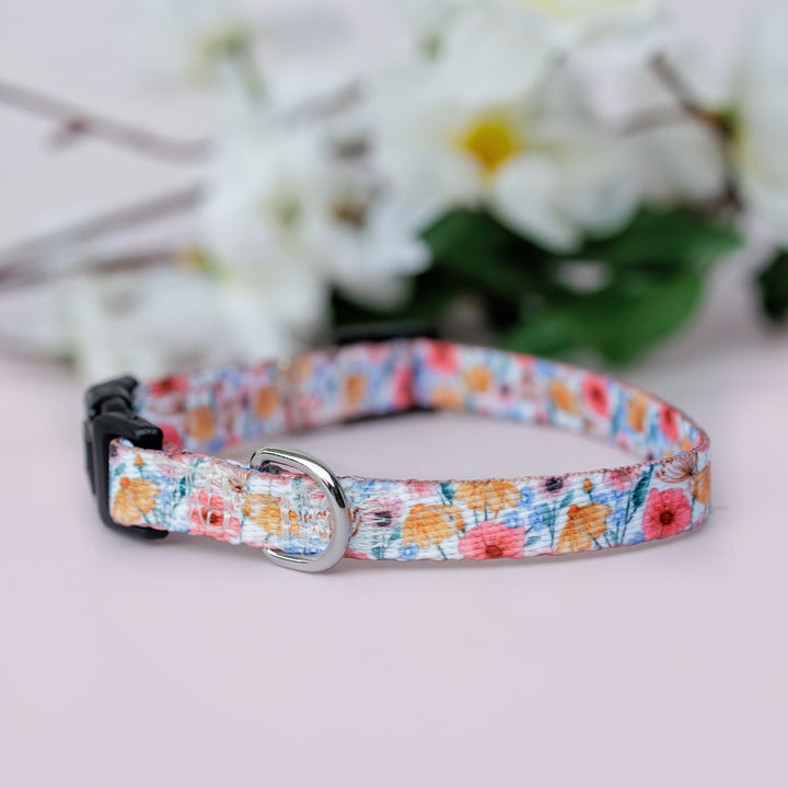 A Peachy Posies Floral pet collar with a floral design in pastel colours, shown from the side with a black buckle and silver D-ring, resting on a light surface with white flowers in the background