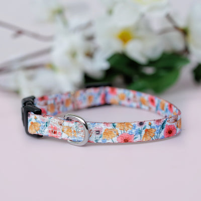 A Peachy Posies Floral pet collar with a floral design in pastel colours, shown from the side with a black buckle and silver D-ring, resting on a light surface with white flowers in the background