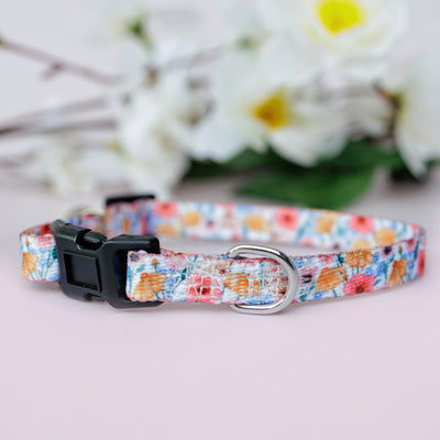 Side view of a Peachy Posies Floral pet collar, showcasing the floral pattern and sturdy black buckle, with silver D-rings on both ends, placed on a light surface with blurred flowers behind