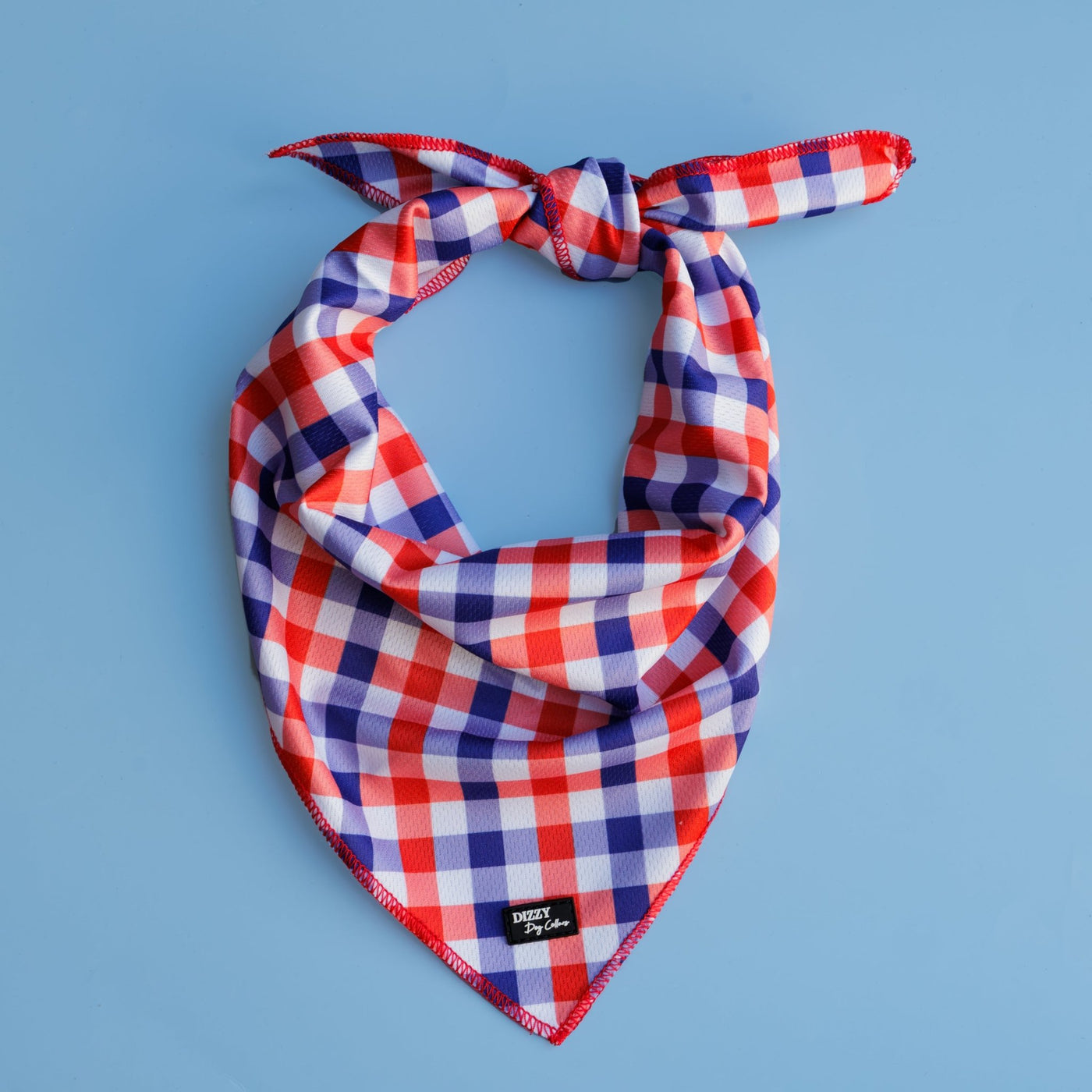 A red, white, and blue gingham-patterned dog bandana with a tied knot, displayed flat on a light blue background. The Dizzy Dog Collars logo is visible on the bottom edge of the bandana