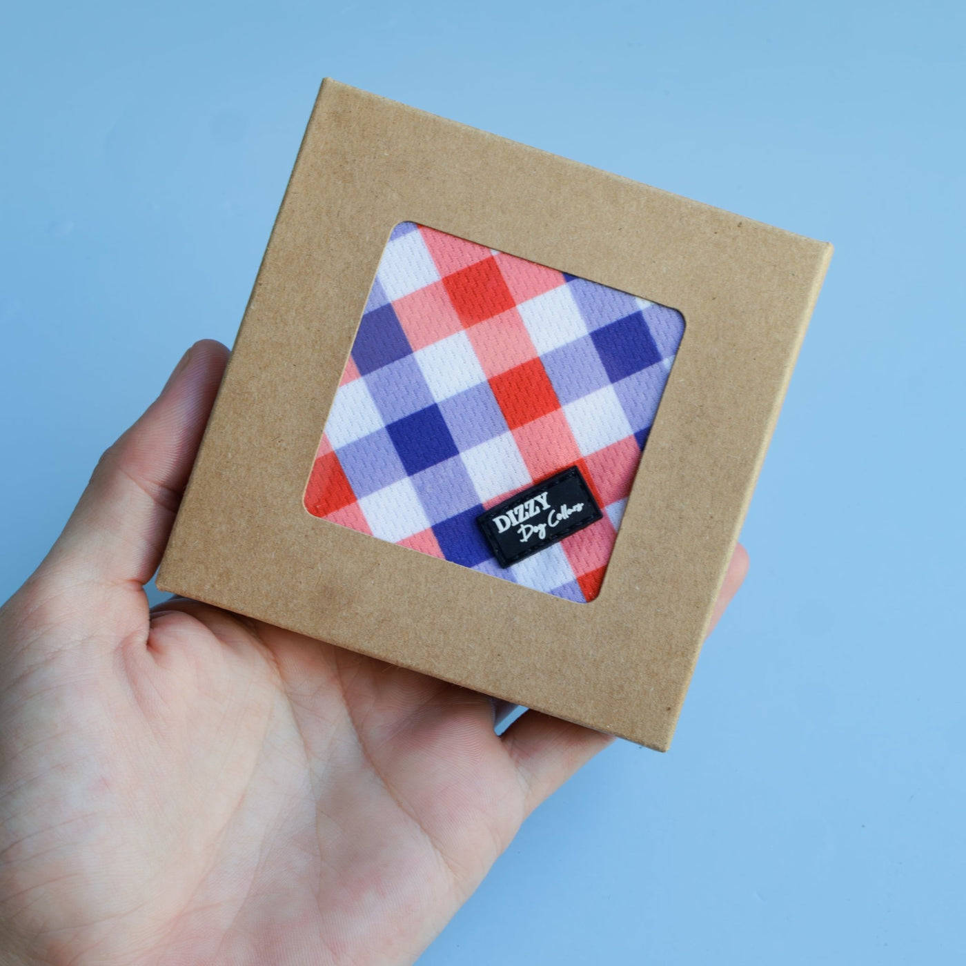 A red, white, and blue gingham-patterned dog bandana in a brown cardboard display box with a clear window, showcasing the Dizzy Dog Collars logo. The bandana is being held in a person's hand against a light blue background