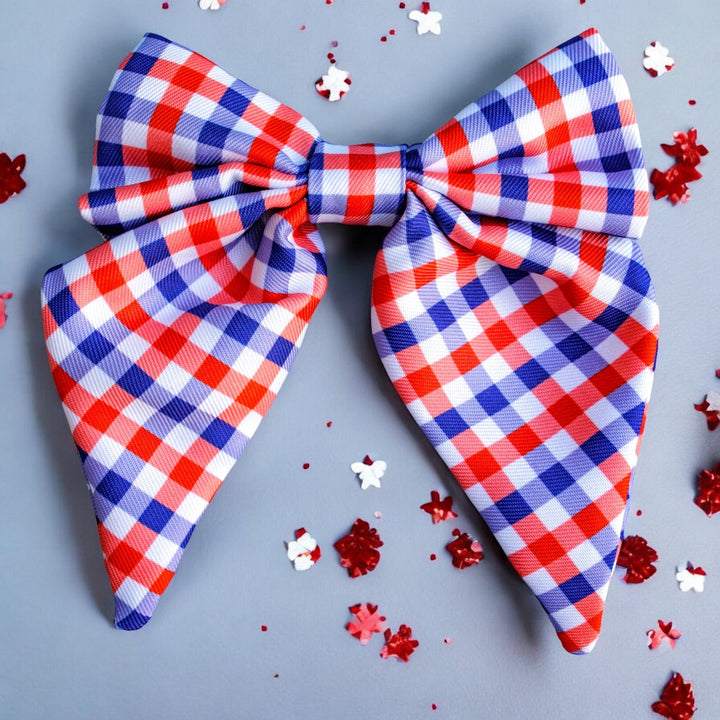  large red, white, and blue gingham-patterned bow tie with pointed ends, displayed on a light blue background. Red, white, and blue circular confetti surrounds the bow, with soft shadows falling across the scene